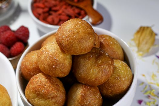 Petits beignets dorés et moelleux empilés dans un bol, accompagnés de fruits rouges en arrière-plan.
